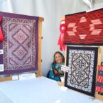 Barbara Teller Ornelas with a large tapestry at left. The contemporary designs at the right are by her son, Michael Ornelas. The Two Grey Hills double diamond piece is by Barbara.